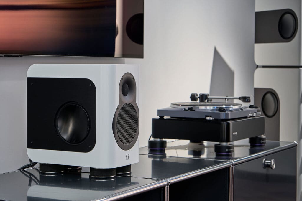 A SEVEN speaker stands on hi-fi furniture beside a turntable, with additional speakers in the background.